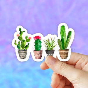 May include: Four watercolor cactus stickers in pots with different designs. The stickers are on a white background and are being held by a hand.
