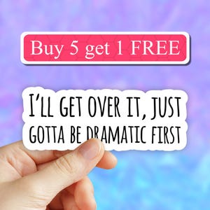 May include: White sticker with black text that reads "I'LL GET OVER IT, JUST GOTTA BE DRAMATIC FIRST". A hand is holding the sticker. A pink and blue background with the text "Buy 5 get 1 FREE" in a red box is visible in the background.