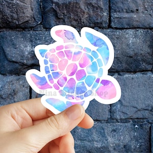 May include: A watercolor-style sticker of a sea turtle in shades of pink, purple, and blue. The turtle has a white outline and is held by a hand. The sticker has the text "Nina Shop".