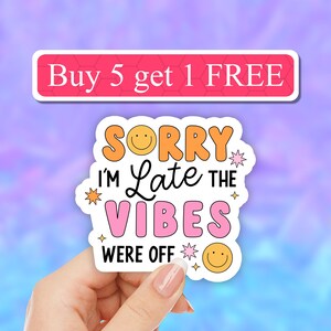 May include: A white sticker with the text "Sorry I'm Late The Vibes Were Off" in pink, orange, and yellow with smiley faces and stars. The sticker is being held by a hand.