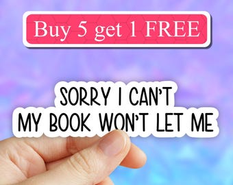 May include: White sticker with black text that reads "SORRY I CAN'T MY BOOK WON'T LET ME". A hand is holding the sticker. A pink and blue background with the text "Buy 5 get 1 FREE" in a red banner is visible in the background.
