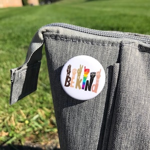 May include: A white button with the words "BE KIND" in black letters. The letters are formed by different colored hands in various hand gestures. A rainbow stripe is incorporated into the design.