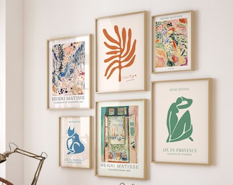 May include: A collection of framed art prints featuring the work of Henri Matisse. The prints are displayed on a white wall above a wooden desk with a lamp, books, and potted plants. The prints include a landscape, a still life, a portrait, and a cat.