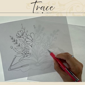 May include: A sheet of white paper with a black line drawing of a book and flowers, partially covered by a translucent sheet of tracing paper. A pink mechanical pencil is used to trace the floral design onto the semi-transparent overlay.