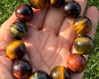 May include: A beaded bracelet featuring polished, spherical gemstones in varying shades of golden-brown, deep red, and dark blue. The beads exhibit a chatoyant, silky lustre characteristic of tiger's eye, arranged in a circular pattern on an elastic cord.