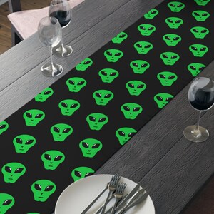May include: A black table runner featuring a repeating pattern of green alien faces. The runner is placed on a dark wooden table set for a meal, with plates, silverware, and wine glasses. A bowl of fruit sits at the table's center.