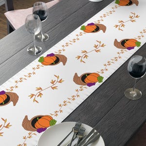 May include: White table runner with a repeating pattern of a cornucopia, pumpkins, and grapes. The runner is decorated with brown and orange autumn leaves. The table is set with plates, silverware, and wine glasses.