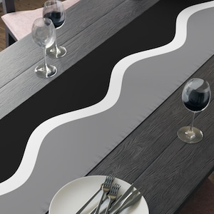 May include: A black wooden dining table with a decorative table runner. The runner features a wavy design with black, gray, and white stripes. The table is set with wine glasses, plates, and silverware, and a bowl of fruit.