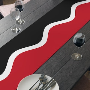 May include: A black dining table with a red and black table runner. The runner has a white wavy design separating the red and black sections. A white plate with silverware and wine glasses are on the table. A bowl of fruit sits at the end.