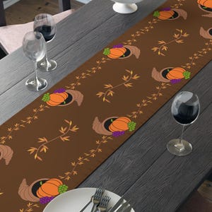 May include: A brown table runner with a repeating pattern of pumpkins, grapes, and cornucopias. The runner is placed on a dark wooden table set for a meal, with plates, silverware, and wine glasses.