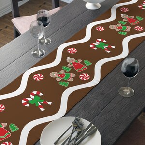 May include: A brown Christmas table runner with a white wavy border and gingerbread man and candy cane designs. The runner is placed on a dark wooden table, set for a holiday meal.