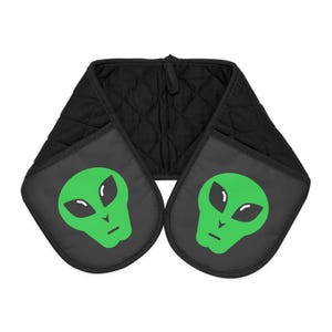 May include: Black quilted oven mitt with two pockets featuring a green alien face design. The alien has large black eyes and a small mouth. The mitt is designed to protect hands from heat when handling hot cookware.