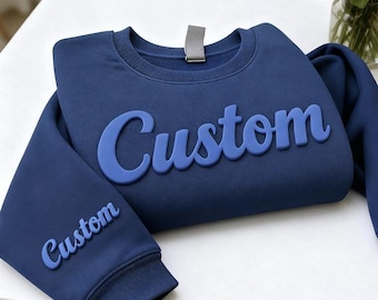 May include: A navy blue sweatshirt with the word "Custom" in a lighter blue, raised font across the chest. The word "Custom" is also on the left sleeve. The sweatshirt is folded on a white surface.