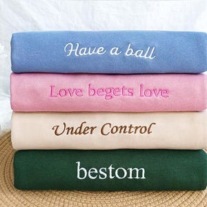 Peut inclure: Quatre échantillons de tissu pliés avec du texte brodé. L'échantillon du haut est bleu clair avec le texte "Have a ball". Le deuxième échantillon est rose avec le texte "Love begets love". Le troisième échantillon est crème avec le texte "Under Control". L'échantillon du bas est vert foncé avec le texte "bestom".