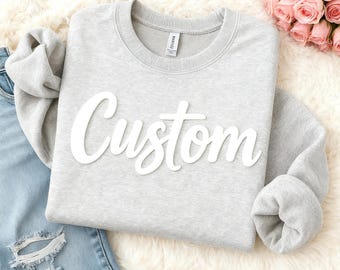 May include: A pale grey sweatshirt with the word "Custom" in white script across the front. The sweatshirt is displayed on a white fluffy surface next to a pair of blue jeans and a bouquet of pink roses.