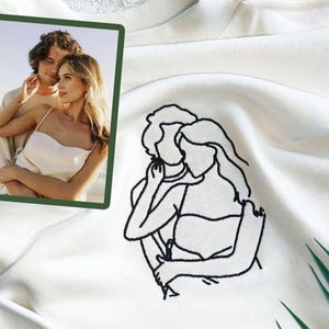May include: A white sweatshirt with a black line drawing of a couple embracing. The drawing is embroidered on the front of the sweatshirt. A photo of a couple is in the background.