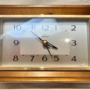 May include: A rectangular wooden clock with a light-colored face. The clock face has black numbers and hands, with the brand name "Sunbeam" visible. The clock is encased in a brown wooden frame.
