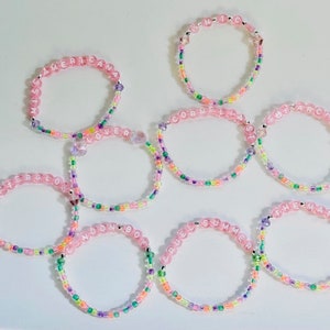 May include: Seven pink beaded bracelets with colorful beads and white lettering spelling out the words "Americana", "Lover", "Afterglow", "London Boy", "Cornelia Street", "The Man", and "Cruel Summer".