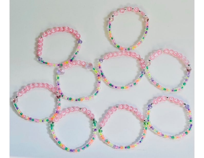 May include: Seven pink beaded bracelets with colorful beads and white lettering spelling out the words "Americana", "Lover", "Afterglow", "London Boy", "Cornelia Street", "The Man", and "Cruel Summer".