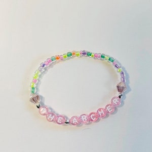 May include: A pink beaded bracelet with the word "THE ARCHER" spelled out in white letters. The beads are interspersed with small, clear, iridescent beads.