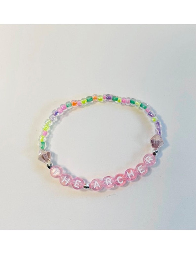 May include: A pink beaded bracelet with the word "THE ARCHER" spelled out in white letters. The beads are interspersed with small, clear, iridescent beads.