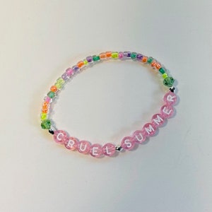 May include: A pink and green beaded bracelet with the words "Cruel Summer" spelled out in white letters.