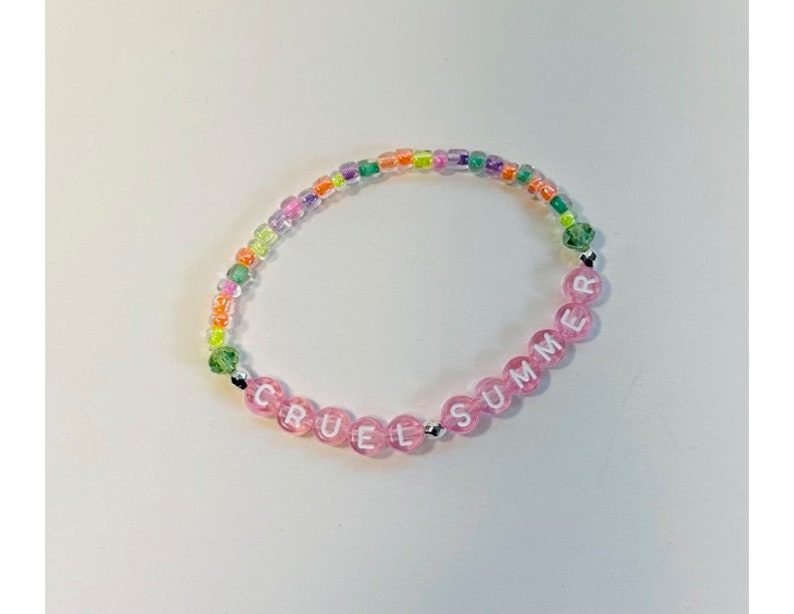 May include: A pink and green beaded bracelet with the words "Cruel Summer" spelled out in white letters.