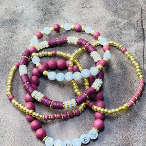May include: A set of maroon and gold beaded bracelets with white beads that spell out "AGGIES" and "BIG" with a heart in between.