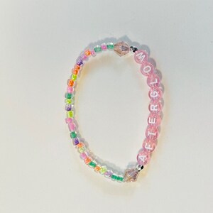May include: A pink beaded bracelet with the word "AFTERGLOW" spelled out in white letters. The beads are interspersed with small, iridescent, multi-colored beads.