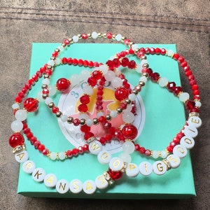 May include: Four red and white beaded bracelets with gold accents. The bracelets spell out "Arkansas", "Woo", "Pig", and "Sooie".
