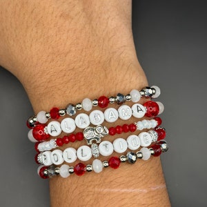 May include: A set of five bracelets featuring red and white beads, silver beads, and white letter beads spelling "ROLL TIDE" and "ALABAMA". A silver elephant charm is attached to one of the bracelets.