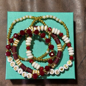 May include: A set of five bracelets with maroon, white, and gold beads. The bracelets spell out "AGGIES" and "SIG EM" with a heart symbol between the words.
