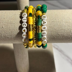 May include: A set of five bracelets with alternating yellow and green beads. The bracelets spell out "SIC 'EM" and "BAYLOR".
