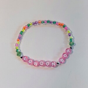 May include: A pink beaded bracelet with the word "LONDON BOY" spelled out in white letters. The bracelet is made with clear, iridescent beads in shades of green, purple, and orange.