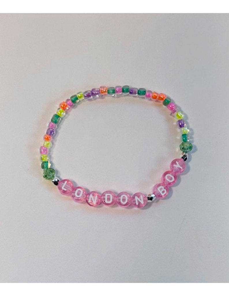 May include: A pink beaded bracelet with the word "LONDON BOY" spelled out in white letters. The bracelet is made with clear, iridescent beads in shades of green, purple, and orange.