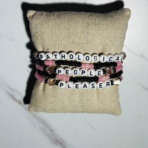May include: Four beaded bracelets with the words "Pathological", "People", and "Pleaser" spelled out in white letters on black beads. The bracelets are stacked on top of each other on a beige fabric surface.