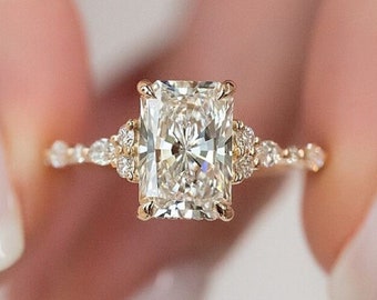 Puede incluir: Un anillo de compromiso de oro con una gran piedra central de diamante rectangular y transparente. La banda está adornada con diamantes más pequeños.