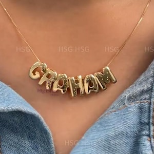 以下が含まれることがあります： 「GRAHAM」という名前が個々の文字で綴られたゴールドカラーのネックレス。デニムジャケットの上から着用されています。文字はブロック体で、繊細なチェーンで繋がれています。「HSG」の文字も表示されています。