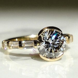 May include: A gold ring featuring a large, round, clear gemstone. The band is adorned with smaller, rectangular gemstones. The ring is set against a white surface, with soft lighting.