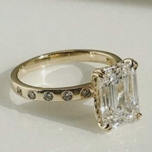 Puede incluir: Un anillo de compromiso dorado con un gran diamante de talla esmeralda. La banda presenta pequeños diamantes redondos. El anillo se muestra sobre un fondo blanco, resaltando su diseño elegante.