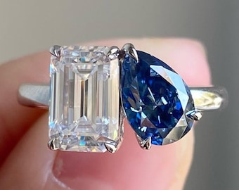 May include: A silver ring featuring two gemstones: a rectangular, clear-cut diamond and a pear-shaped, deep blue gemstone. The ring's design showcases a modern, elegant aesthetic, perfect for engagement or special occasions.