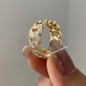 May include: A gold ring with a thick, pave-set diamond band in a chain link design. The ring is on a finger with a light pink nail polish.