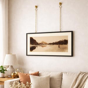 Può includere: Una fotografia di paesaggio in seppia incorniciata, sospesa da catene dorate. La cornice nera racchiude una scena di acqua e montagne. Una lampada bianca e un divano sono visibili in primo piano.