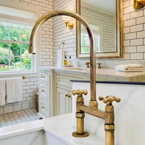 Peut inclure: Gros plan sur un robinet en laiton de style vintage avec des poignées en croix. Le robinet est placé sur un fond de carreaux de métro blancs, d'un comptoir en marbre et d'un grand miroir. La salle de bain a un design classique et élégant.