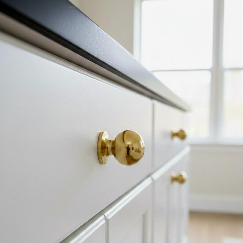 K&ouml;nnte beinhalten: Nahaufnahme von wei&szlig;en Schrank Schubladen mit goldenen kugelf&ouml;rmigen Kn&ouml;pfen. Die Schubladen befinden sich unter einer schwarzen Arbeitsplatte. Die Kn&ouml;pfe sind rund und gl&auml;nzend und reflektieren das Licht. Der Schrank befindet sich in einem hellen Raum mit einem Fenster.