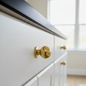 K&ouml;nnte beinhalten: Nahaufnahme von wei&szlig;en Schrank Schubladen mit goldenen kugelf&ouml;rmigen Kn&ouml;pfen. Die Schubladen befinden sich unter einer schwarzen Arbeitsplatte. Die Kn&ouml;pfe sind rund und gl&auml;nzend und reflektieren das Licht. Der Schrank befindet sich in einem hellen Raum mit einem Fenster.