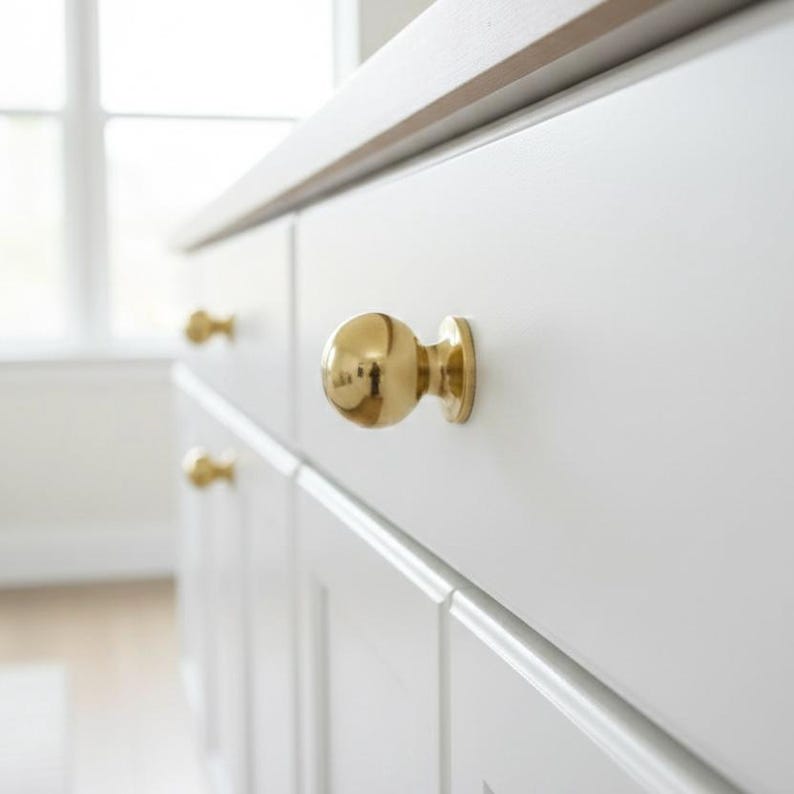 K&ouml;nnte beinhalten: Nahaufnahme eines wei&szlig;en Schranks mit goldfarbenen, kugelf&ouml;rmigen Kn&ouml;pfen. Die Kn&ouml;pfe sind an den Schrankt&uuml;ren befestigt. Der Schrank ist Teil eines gr&ouml;&szlig;eren M&ouml;belst&uuml;cks, m&ouml;glicherweise eines K&uuml;chenschranks oder einer Kommode.