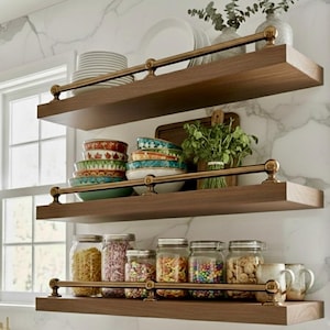 Puede incluir: Estanterías de madera de tres niveles con detalles de latón, que exhiben varios artículos de cocina. El estante superior contiene platos blancos y un jarrón con vegetación. El estante del medio presenta cuencos coloridos y una tabla de cortar de madera. El estante inferior muestra frascos de vidrio llenos de pasta, caramelos y nueces.