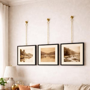 May include: Three framed landscape prints with black frames and cream-coloured mats are suspended from the wall by gold-coloured chains. The prints depict sepia-toned scenes of water and trees. A white lamp with a pleated shade sits on a table below.