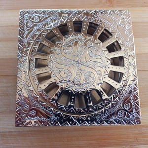 May include: A square, silver drain cover with an intricate, ornate design. The cover has a circular, open center with a decorative pattern.